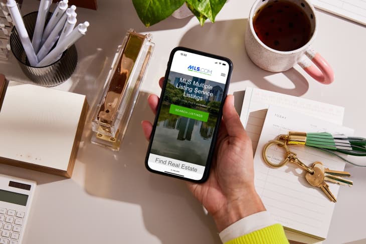 Person holding phone at desk with MLS website pulled up