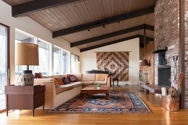 angled ceiling, dark wood paneled ceiling, wood beams, wood floors, blue and coral rug, diamond tapestry, tan tall lamp shade, exposed brick, fireplace, white sectional, wood coffee table, copper bucket, throw pillows