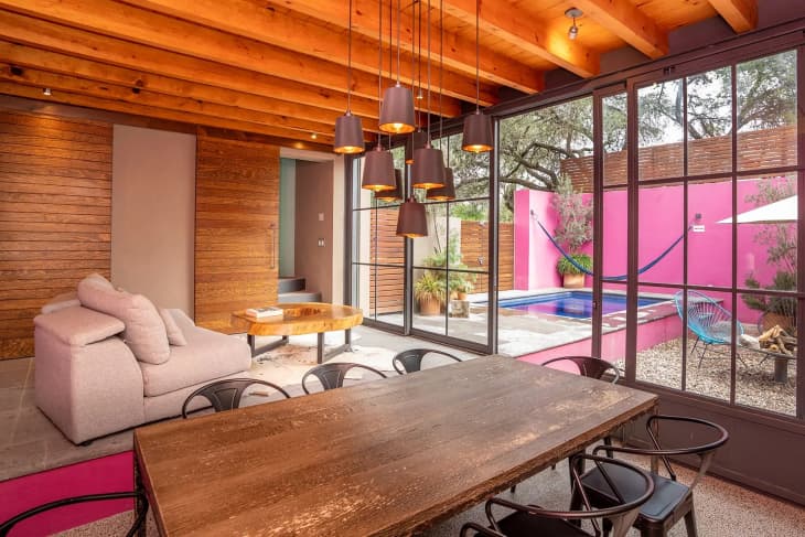 Large window in vacation home with pink exterior wall in gravel lined fire pit. Hanging pendant lamps over wooden dining table and small armchair in dining room.