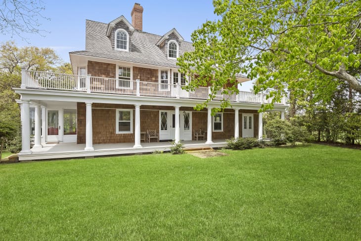Three-story house with shingle siding, white columns, wraparound porch, and lush green lawn surrounded by trees.