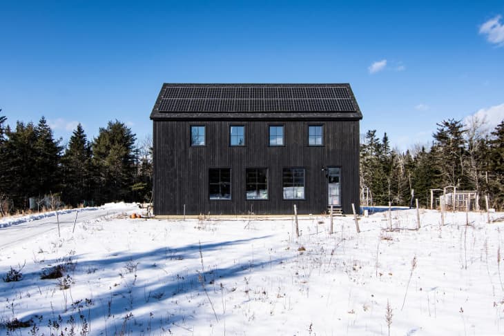 Black modern house with solar panels, surrounded by snow and trees under a clear blue sky.