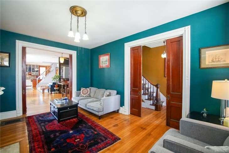 Living room with teal walls, gray sofas, red patterned rug, and wooden doors leading to a staircase and dining area.