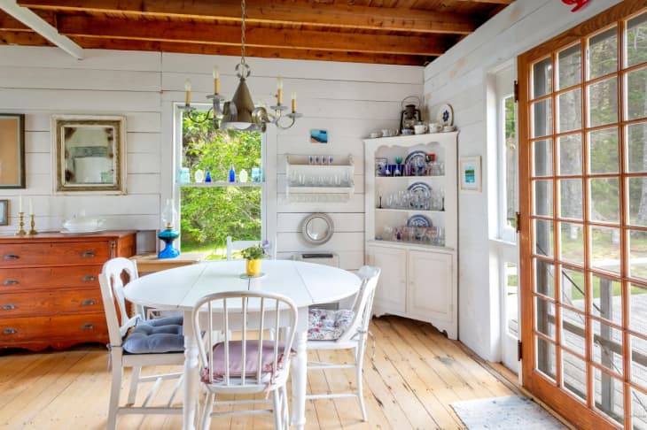 Dining room with white table, wooden chairs, chandelier, and glass cabinet, next to large windowed doors and a wooden dresser.