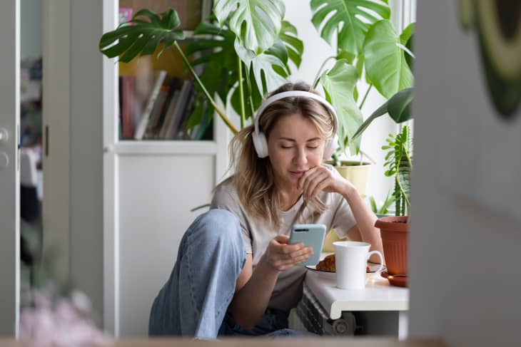 Woman with headphones using smartphone, sitting by a table with coffee and plants.