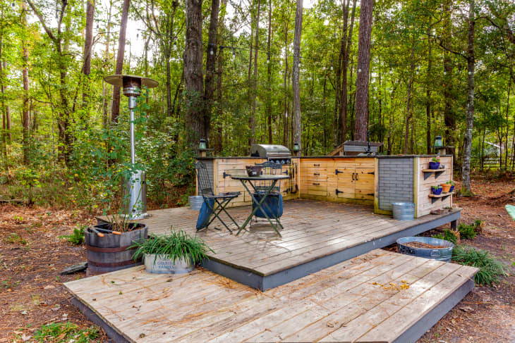 Outdoor wooden deck with a grill, patio heater, folding chairs, and potted plants surrounded by trees.