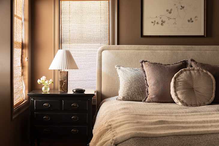 Cozy bedroom featuring a beige upholstered bed, decorative pillows, a black nightstand with a lamp, and a small vase of flowers.