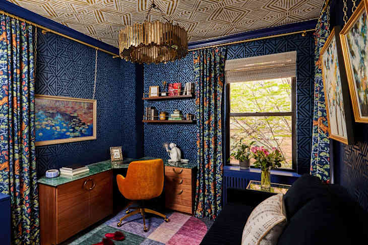 Cozy home office with blue geometric wallpaper, colorful floral curtains, green desk, and a gold chandelier.