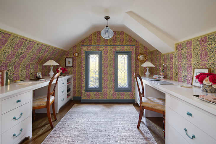 A cozy home office with two white desks, patterned wallpaper, and large windows. Decor includes a lamp, flowers, and books.