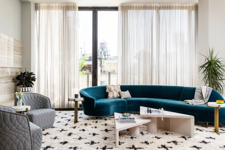 Modern living room with teal curved sofa, gray armchairs, geometric coffee tables, and large windows with sheer curtains.
