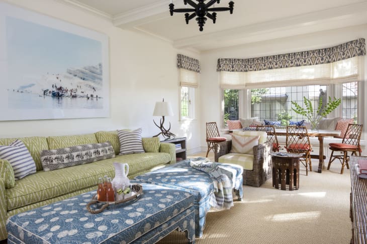 Living room with green sofa, patterned ottoman, wicker chairs, large windows, and a wall art piece.