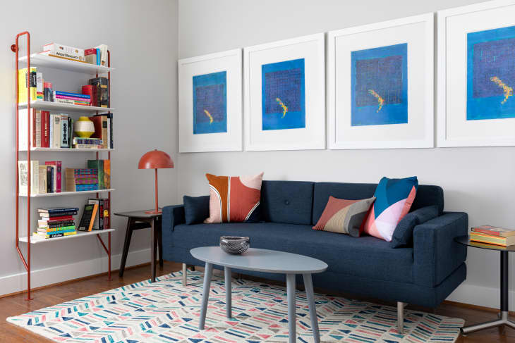 Living room with blue sofa, colorful pillows, geometric rug, bookshelf, and four framed blue artworks on the wall.