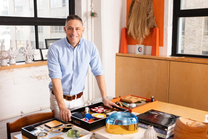 Man in a light blue shirt standing at a table with design objects, books, and decor in a bright room with large windows.