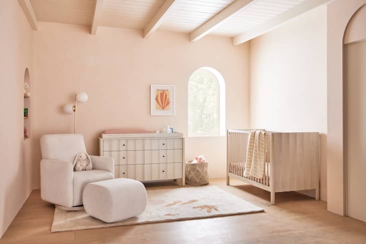 Nursery with a white armchair, ottoman, wooden crib, dresser, and wall art, featuring soft pink walls and a window.
