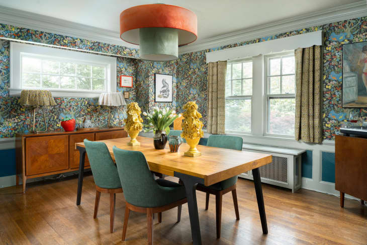Wooden dining table with teal chairs in room with patterned wallpaper and short curtains.