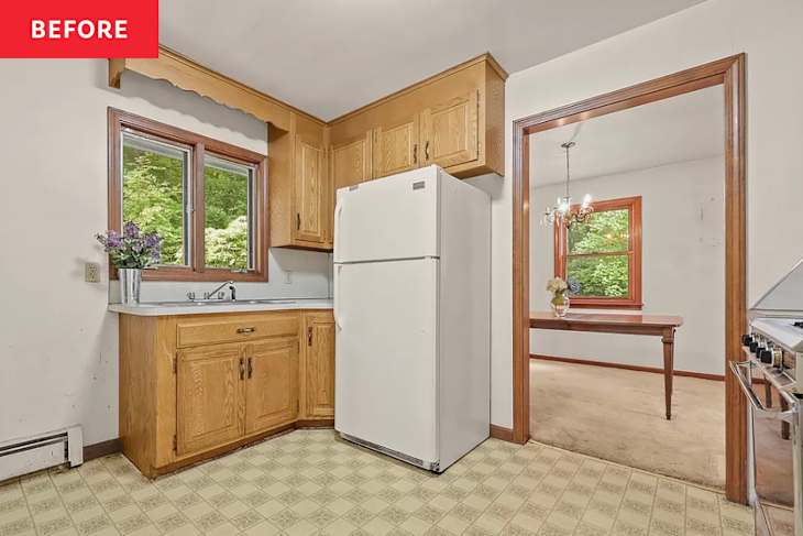 Kitchen with wooden cabinets, white refrigerator, and a window with flowers. Dining area visible through an open doorway.