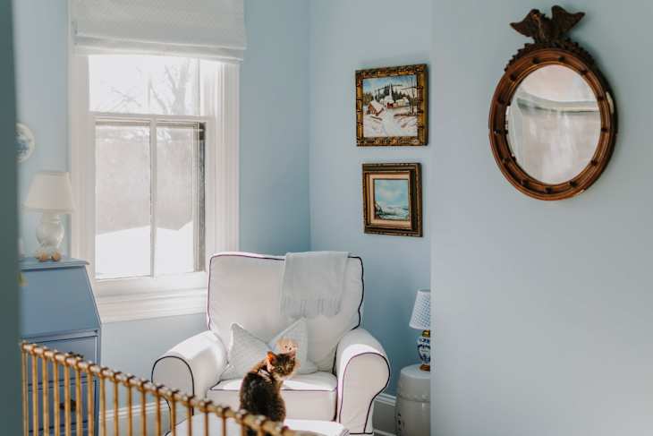 Cozy room with light blue walls, a white armchair, a cat, and framed artwork. A window and lamp are visible.