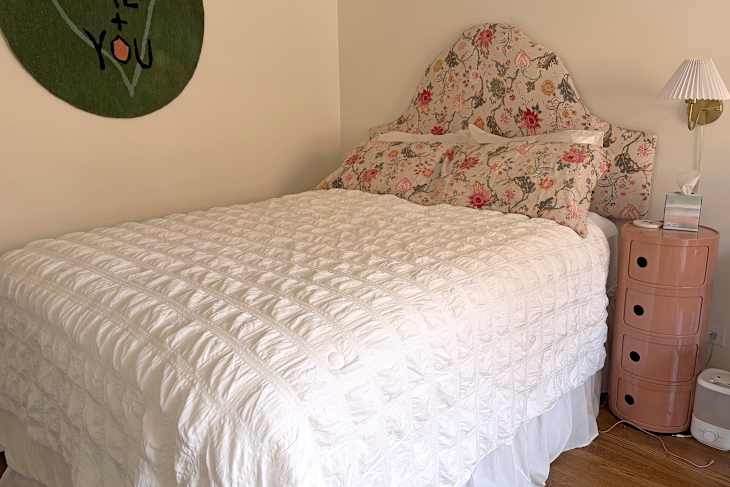 Cozy bedroom featuring a white quilted bedspread, floral pillows, a pink nightstand, and a wall-mounted lamp.