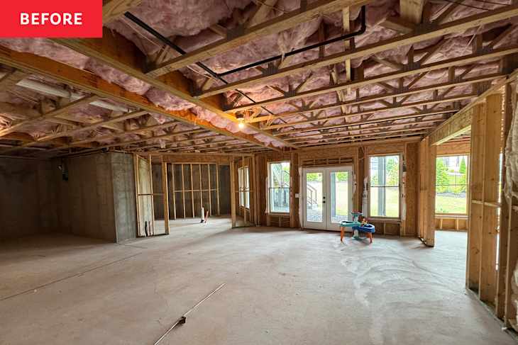 Open space under construction with exposed wooden beams, pink insulation, and concrete flooring. Two large windows and a door lead outside.