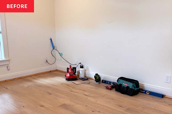 Empty room corner with light wood flooring, tools including a red sander, green laser level, and a black tool bag.