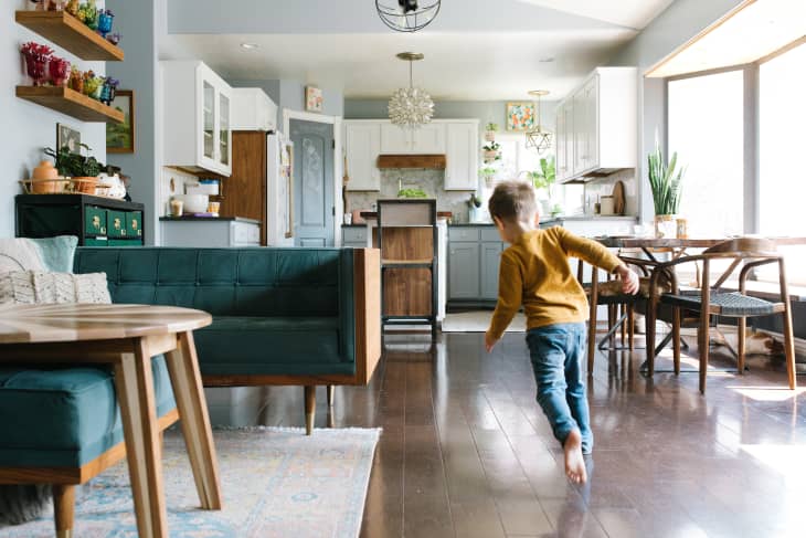 Living space in a home. Photo shows part of a sofa, the dining area, and the kitchen in the background. A young boy is running through the scene.