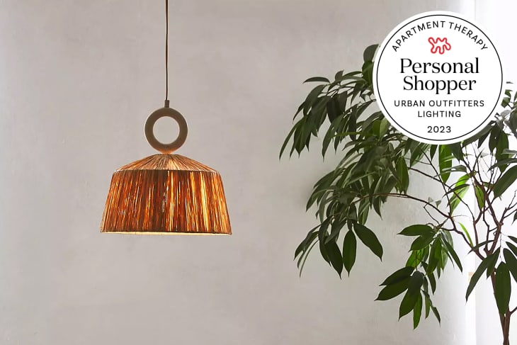 Modern-boho pendant light featuring a resin top ring accenting the woven raffia shade set at an angled slant in a round silhouette. Plant to the right and a seal that reads "Apartment Therapy Personal Shopper: Urban Outfitters Lighting 2023"