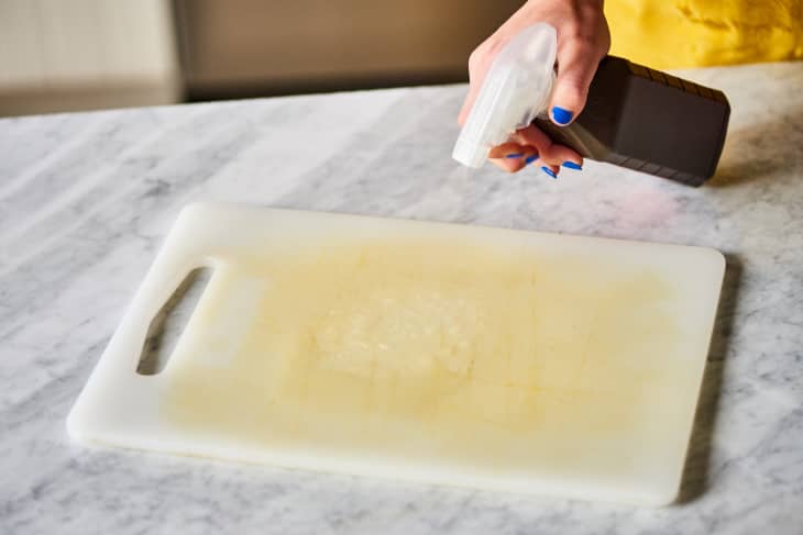Hand spraying cleaner on a stained white cutting board on a marble countertop.