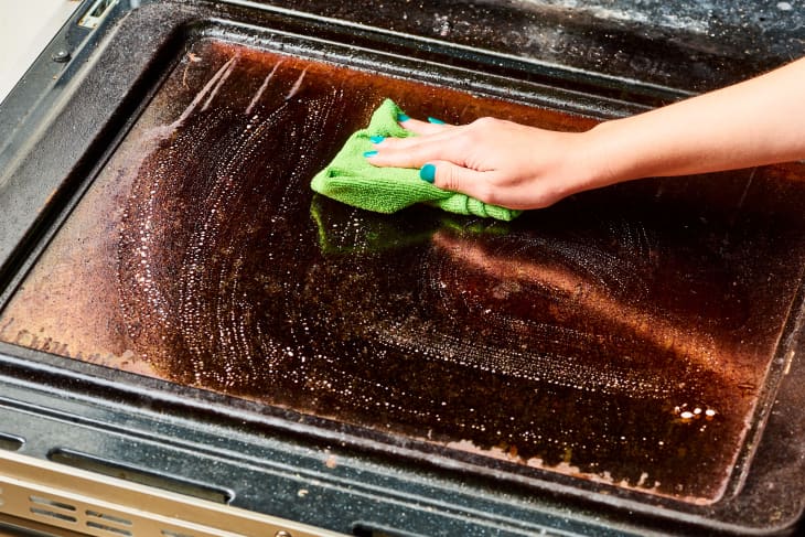 Hand cleaning an oven door with a green cloth and soapy water.