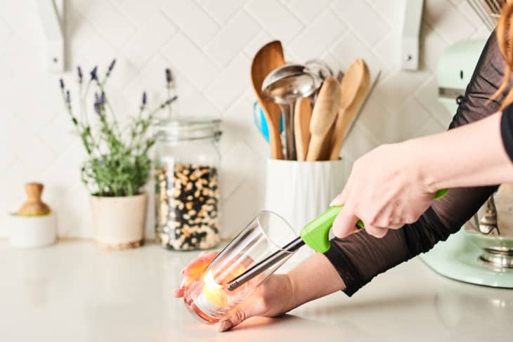 Hand lighting a candle in a glass jar on a kitchen counter with utensils and a plant in the background.
