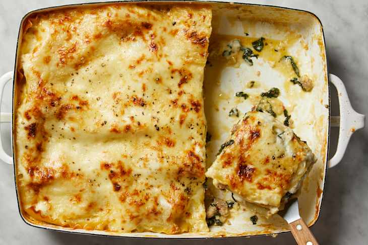 An overhead view of slice of white lasagna being lifted out of a baking dish on a white marble background