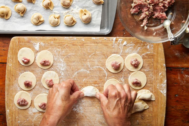 Hands fold a dumpling in the middle of a cutting board lined with dough rounds.