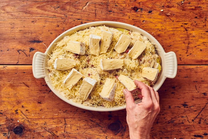 Cut wedges of cheese being placed on top of large circular dish with shredded cheese and tartiflette under it