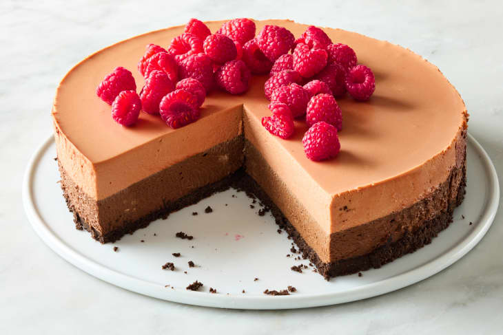 angled shot of triple chocolate mousse cake with a large piece taken out of it, topped with raspberries.