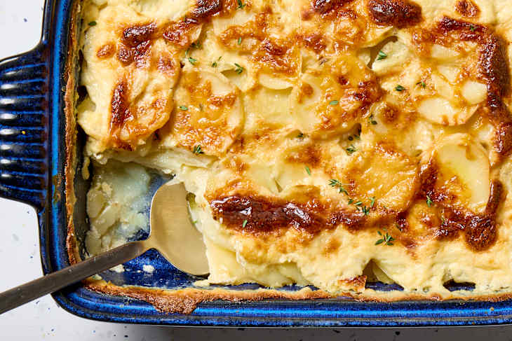 overhead shot of scalloped potatoes in a dark blue baking dish, with a large scoop taken from the bottom left corner.