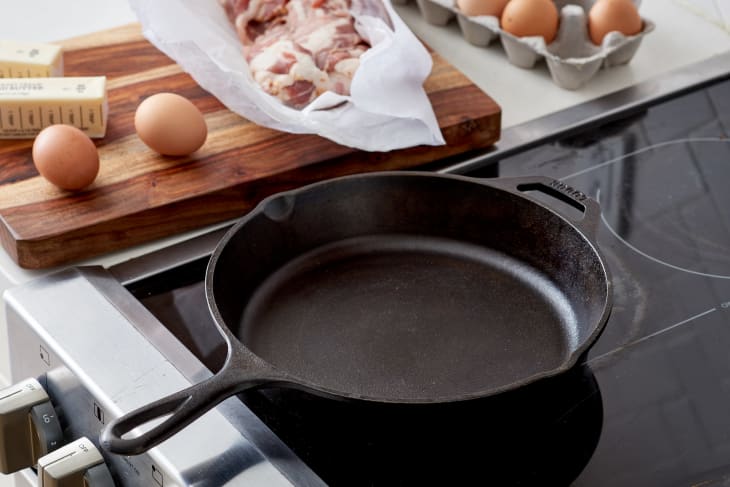A cast iron skillet on top of an electric stove, and besides it is eggs, butter and meat placed on a wooden chopping board