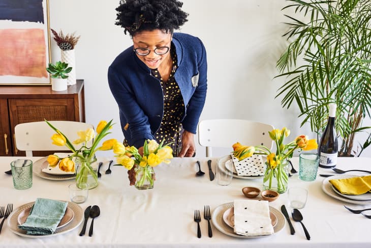 Woman setting a dining table with yellow tulips, plates, and glasses, next to a large plant and a bottle of wine.