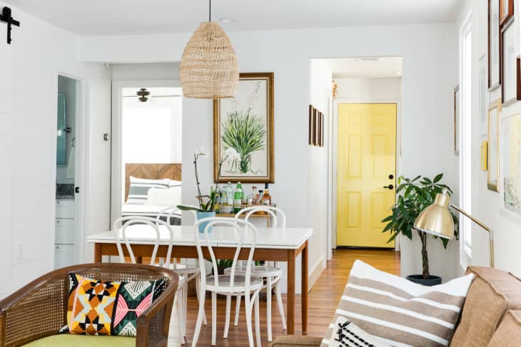 Dining area with white chairs, wooden table, wicker pendant light, and a yellow door in the background.