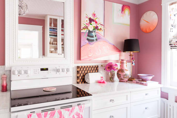 Pink-themed kitchen with white cabinets, floral decor, black lamp, and abstract art on the wall.