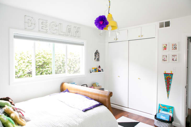 Child's bedroom with a wooden bed, colorful ceiling light, wall art, and books near a window with "DECLAN" above.