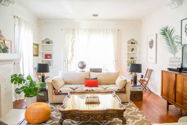 Bright living room with a cream sofa, wooden coffee table, chess set, plants, and decorative shelves.