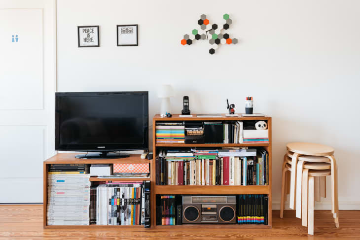 Living room with TV on wooden stand, bookshelves filled with books, stacked stools, and geometric wall art.