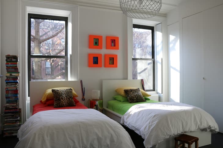Two twin beds with white duvets, colorful pillows, and leopard print cushions in a bright room with two windows and orange wall art.
