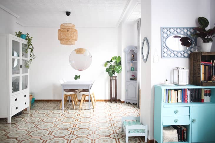 Dining room with white table, wooden chairs, round mirror, wicker pendant light, and plants on patterned tile floor.