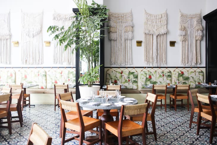 Restaurant interior with macramé wall hangings, patterned floor tiles, wooden chairs, and floral cushioned seating.
