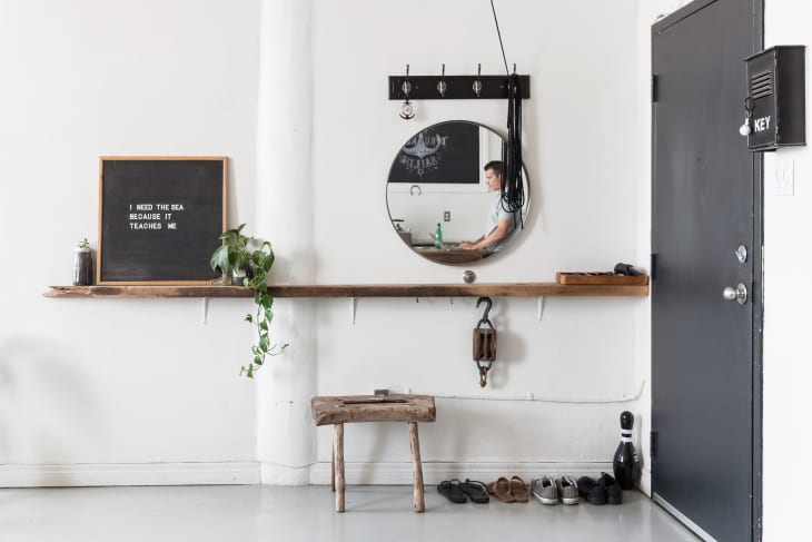 Entryway with a round mirror, wooden shelf, hanging plant, bench, shoes, and a black door with a key box.