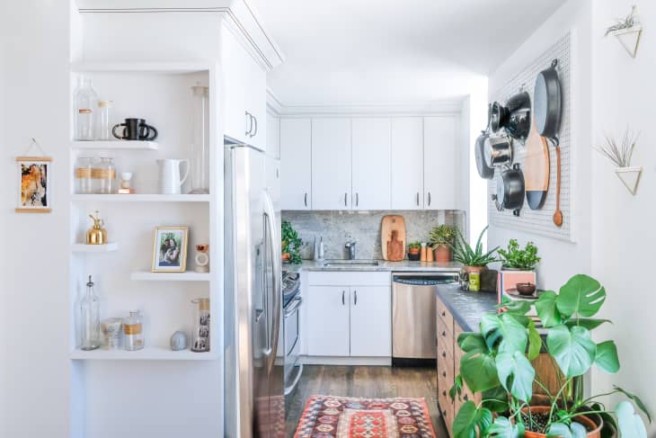 Bright kitchen with white cabinets, open shelving, hanging pots, plants, and a colorful rug on wooden floor.