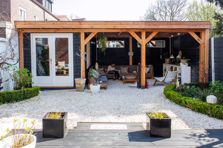 Covered patio with wooden beams, outdoor seating, potted plants, and gravel flooring in a garden setting.