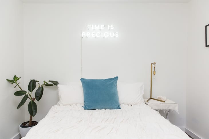 A neon sign that reads "time is precious" above a white bed