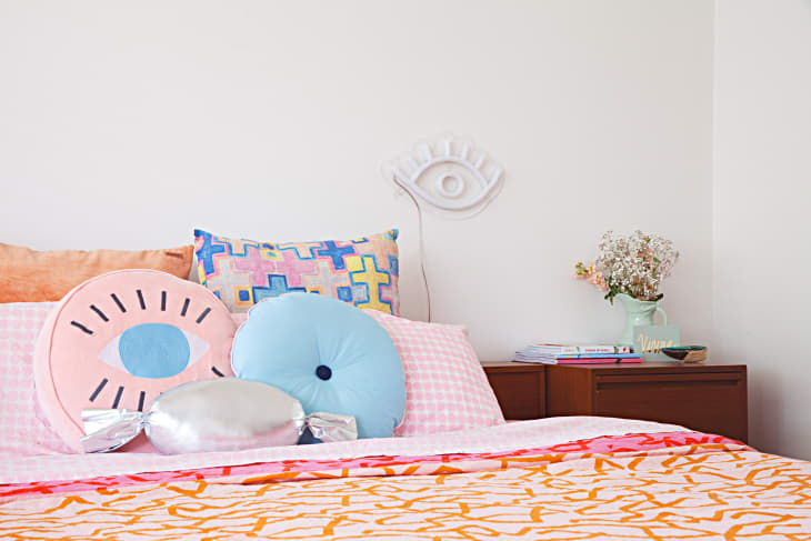 Colorful bedroom with patterned pillows, eye wall art, and a wooden nightstand with books and a vase of flowers.