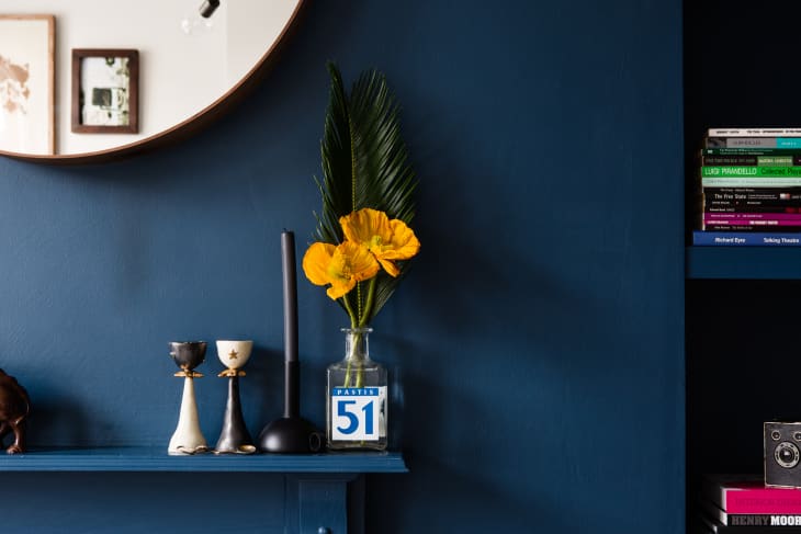 Dark blue wall with a round mirror, yellow flowers in a Pastis 51 bottle, candlesticks, and books on a shelf.