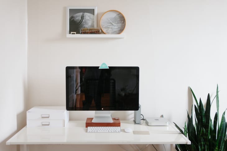 Minimalist desk setup with a computer, keyboard, white storage boxes, and a tall plant. Wall shelf with framed art.